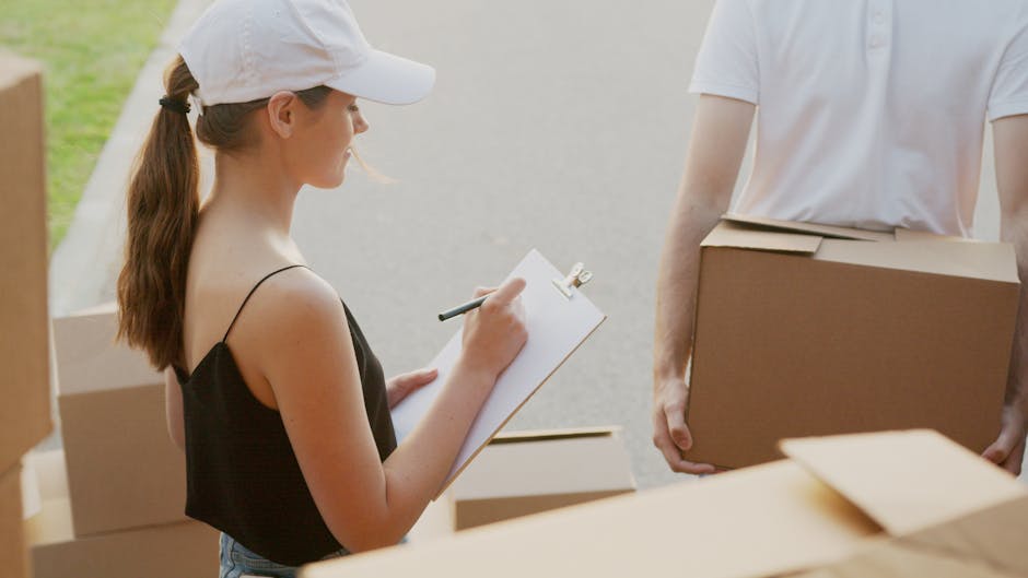 A woman wearing a white cap and black sleeveless top is standing outside on a paved area, holding a clipboard and pen while taking notes during a home relocation process. Several cardboard boxes, some