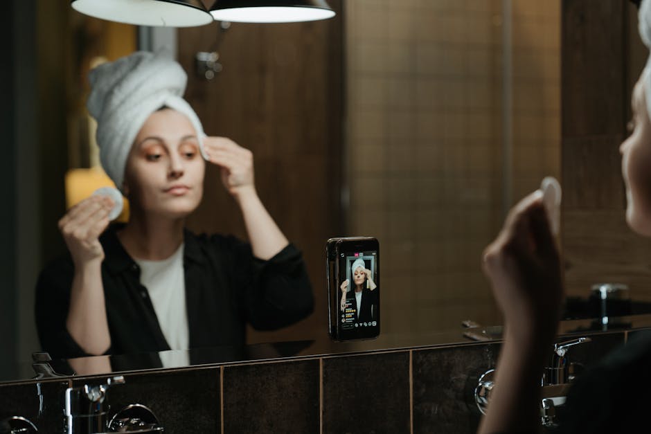 A woman with a towel wrapped around her head is standing in front of a bathroom mirror, applying skincare or makeup with a sponge in one hand while holding another sponge near her temple. The mirror is mounted on a wooden wall panel, and the bathroom countertop beneath it holds a smartphone positioned on a stand, showing her reflection on the screen. The lighting appears soft and warm, illuminating her face and the surrounding area. The scene captures a personal grooming routine during a home relocation or packing process, illustrating the importance of preparing personal belongings like toiletries and skincare items for professional removals. The overall setting emphasizes the indoor environment associated with house move preparations, with focus on interior details such as the mirror, countertop, and everyday grooming items, supported by the professional services of Man with Van Beckenham for home removals and furniture transport.